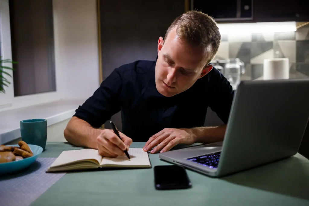 Profissional fazendo anotações de phrasal verbs enquanto estuda no laptop